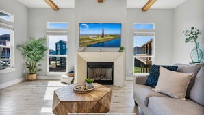 A spacious living room with a fireplace, a large flat-screen TV, and a view of a blue house through the windows. - Perfect Beach Getaway Home!  Beautiful property! (Bolivar Peninsula)