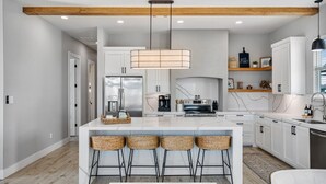 A modern kitchen with white cabinets, a large island with bar stools, a hanging pendant light, and a wooden ceiling beam. - Perfect Beach Getaway Home!  Beautiful property! (Bolivar Peninsula)