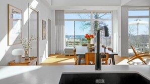 A modern kitchen with a large window, a dining area with a table and chairs, and a view of the sea.