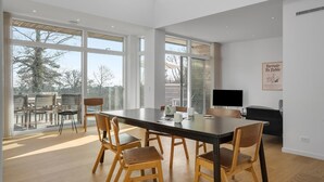A spacious dining area with a long table and wooden chairs, large windows offering a view of the outdoors, and a flat-screen TV mounted on the wall.