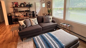 Un salon moderne avec un canapé gris foncé, une table basse et un tapis à motifs. On y trouve également un ventilateur de plafond, une bibliothèque et une fenêtre avec des stores.