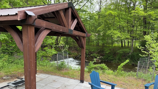 Un belvédère en bois, doté d’une balustrade en métal, surplombe une rivière et est entouré de verdure.