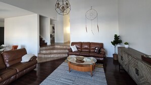 Een ruime woonkamer met een bruine leren bank, een glazen salontafel en een vloerkleed met een patroon. Een trap leidt naar een ander niveau, en een dromenvanger hangt aan de muur.