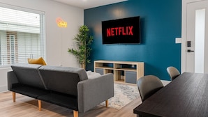 A modern living room with a blue accent wall, a mounted TV displaying 'NETFLIX', a grey sofa, a small wooden table, and a potted plant. - Modern Stylish Oasis in Downtown Dallas w Parking (Dallas)