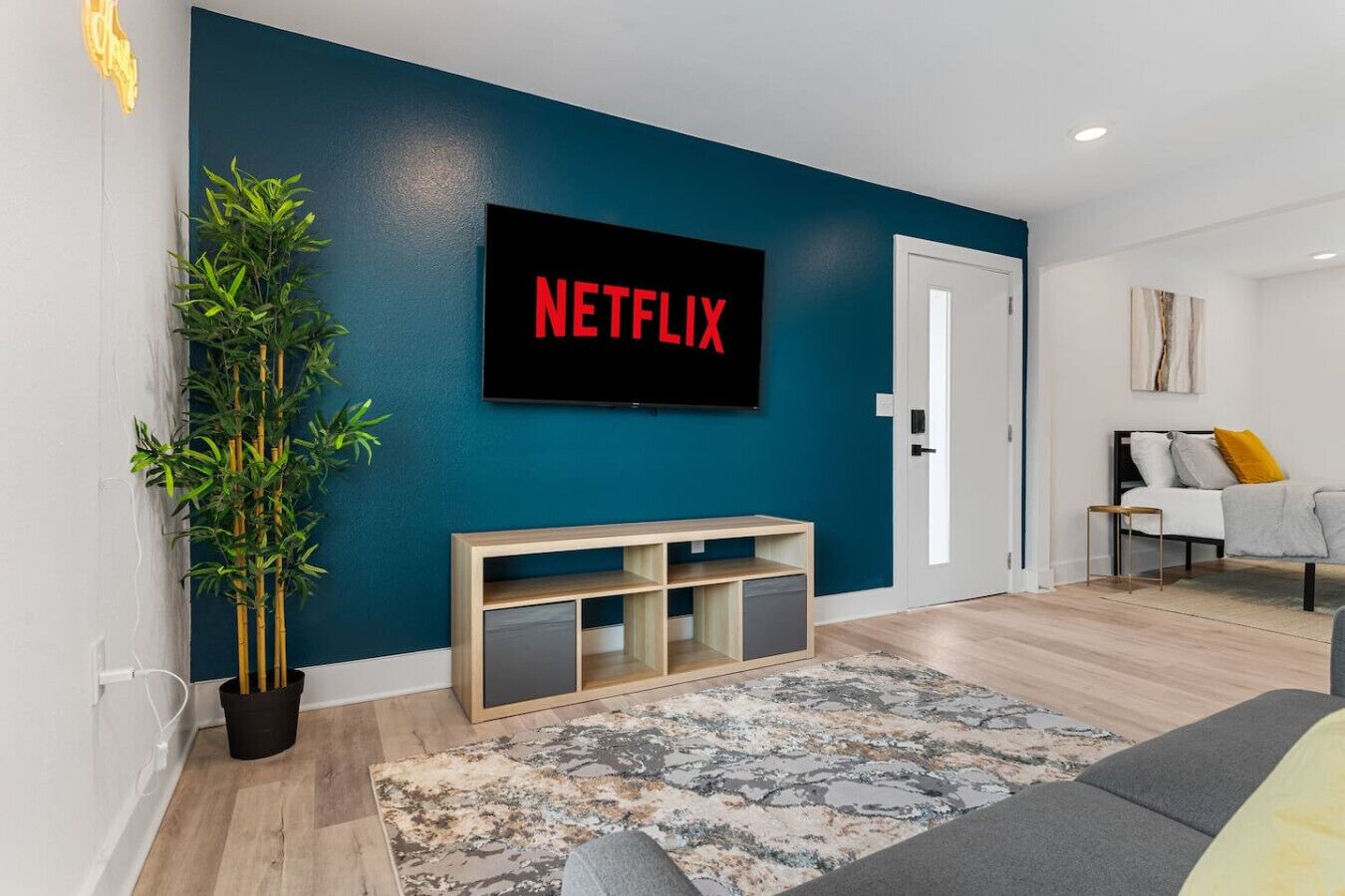 A modern living room with a large TV displaying 'NETFLIX' on the wall, a wooden console table, a patterned rug, and a sofa with cushions.