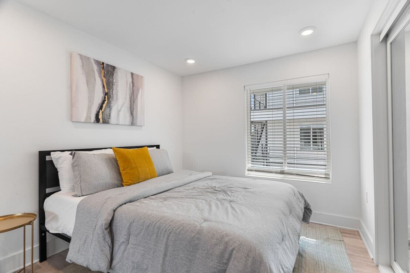 A modern bedroom with a bed, a window with blinds, a painting on the wall, and a small table.