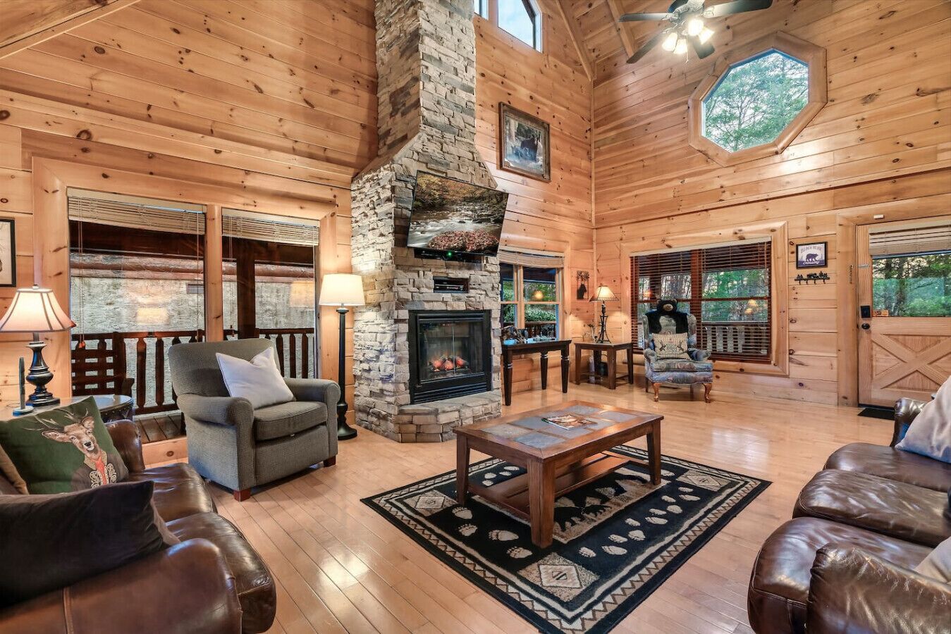 A log cabin interior with a stone fireplace, leather furniture, and large windows.
