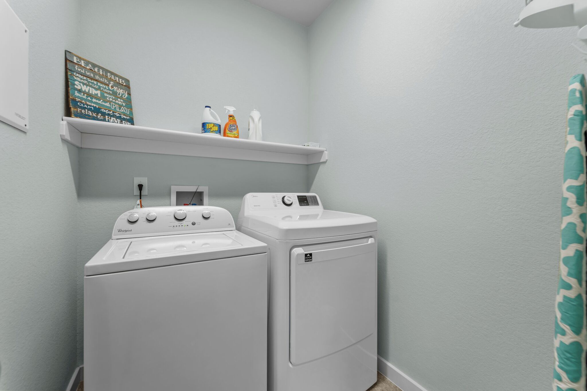 A laundry room with two white Whirlpool washers and a shelf above them holding cleaning supplies.