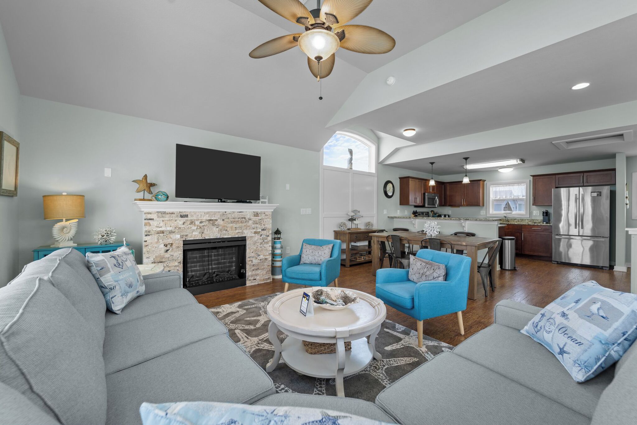 A modern living room with a fireplace, blue armchairs, a dining area, and a kitchen with stainless steel appliances.