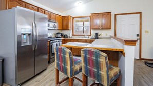 Una cocina con gabinetes de madera, un refrigerador de acero inoxidable, un microondas y una estufa. Hay dos sillas con estampado de cuadros en la isla de la cocina.