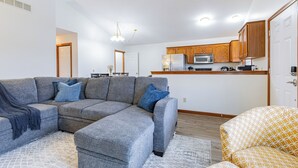 Una sala moderna con un sofá modular, un sillón estampado y una cocina con armarios de madera y un microondas.