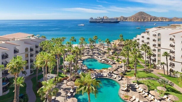 A resort with a swimming pool, palm trees, and a view of the ocean and a cruise ship.
