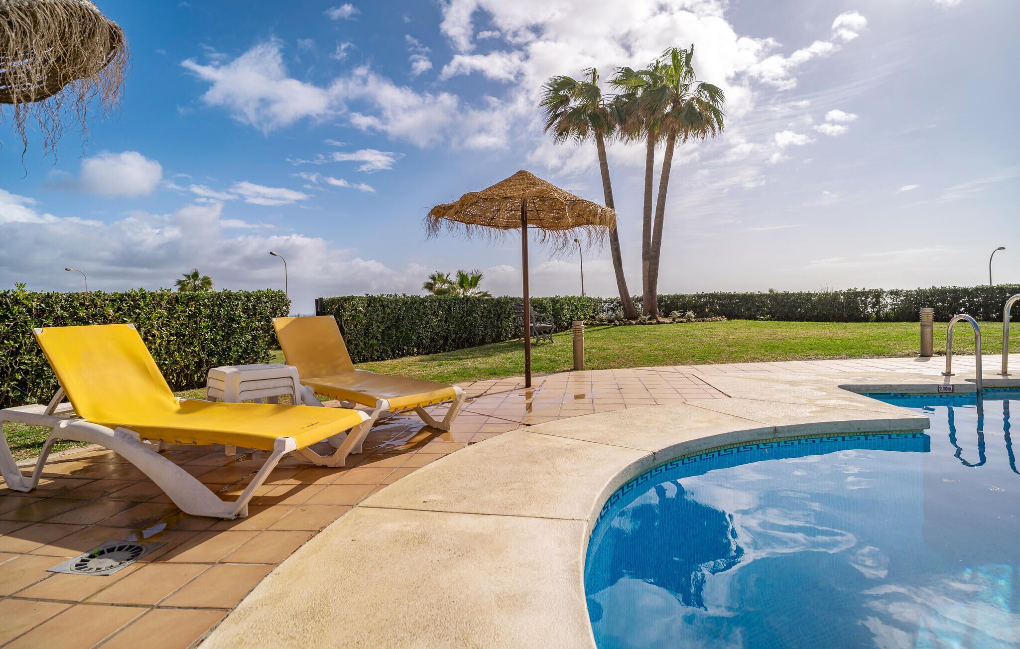 A swimming pool area with yellow lounge chairs, a thatched umbrella, and palm trees.