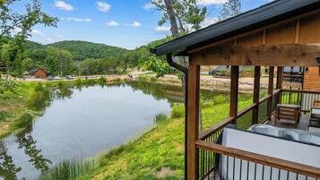 A wooden deck with a hot tub, overlooking a calm lake with a forested hill in the background.