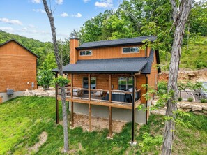 A two-story wooden cabin with a balcony, surrounded by trees and a grassy yard. - Cabin with Pond View, Private Hot Tub, Indoor Pools & Free Attraction Tickets (Hollister)