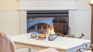 Uma lareira com mantelo branco, uma mesa com um prato de muffins e duas canecas com o logotipo de uma casa.