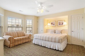 A bedroom with a bed, a sofa, a ceiling fan, and two windows with blinds.