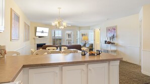 Una cocina amplia con una isla grande, muebles blancos y un candelabro. Hay un comedor con sillas y una mesa, y una sala de estar con un sofá y un televisor.