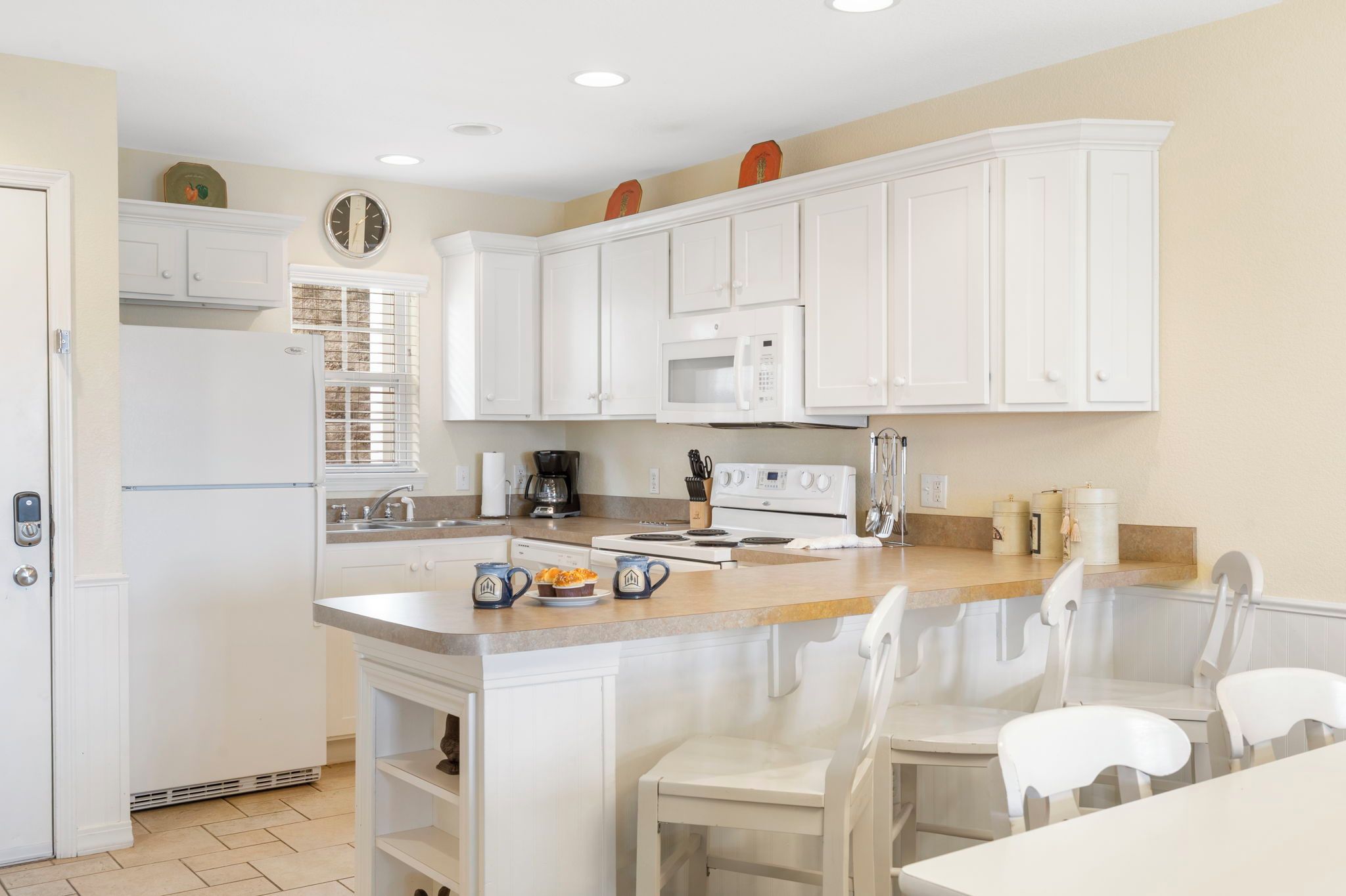 A kitchen with white cabinets, a microwave, a stove, and a sink. There is a window with blinds, a coffee maker, and a pitcher on the counter.