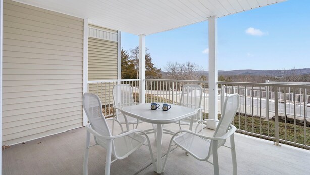 Un balcon abrité avec une table et des chaises blanches, deux tasses sur la table, et une vue sur un parking et des collines en arrière-plan.