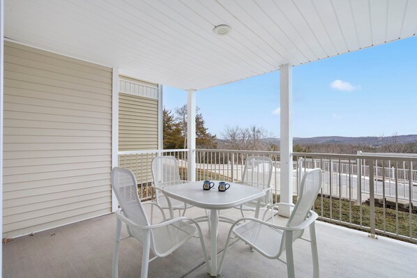 A covered balcony with a white table and chairs, two mugs on the table, and a view of a parking lot and hills in the background.