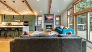 A modern living room with a fireplace, a large window, and a kitchen area with green cabinets and bar stools.