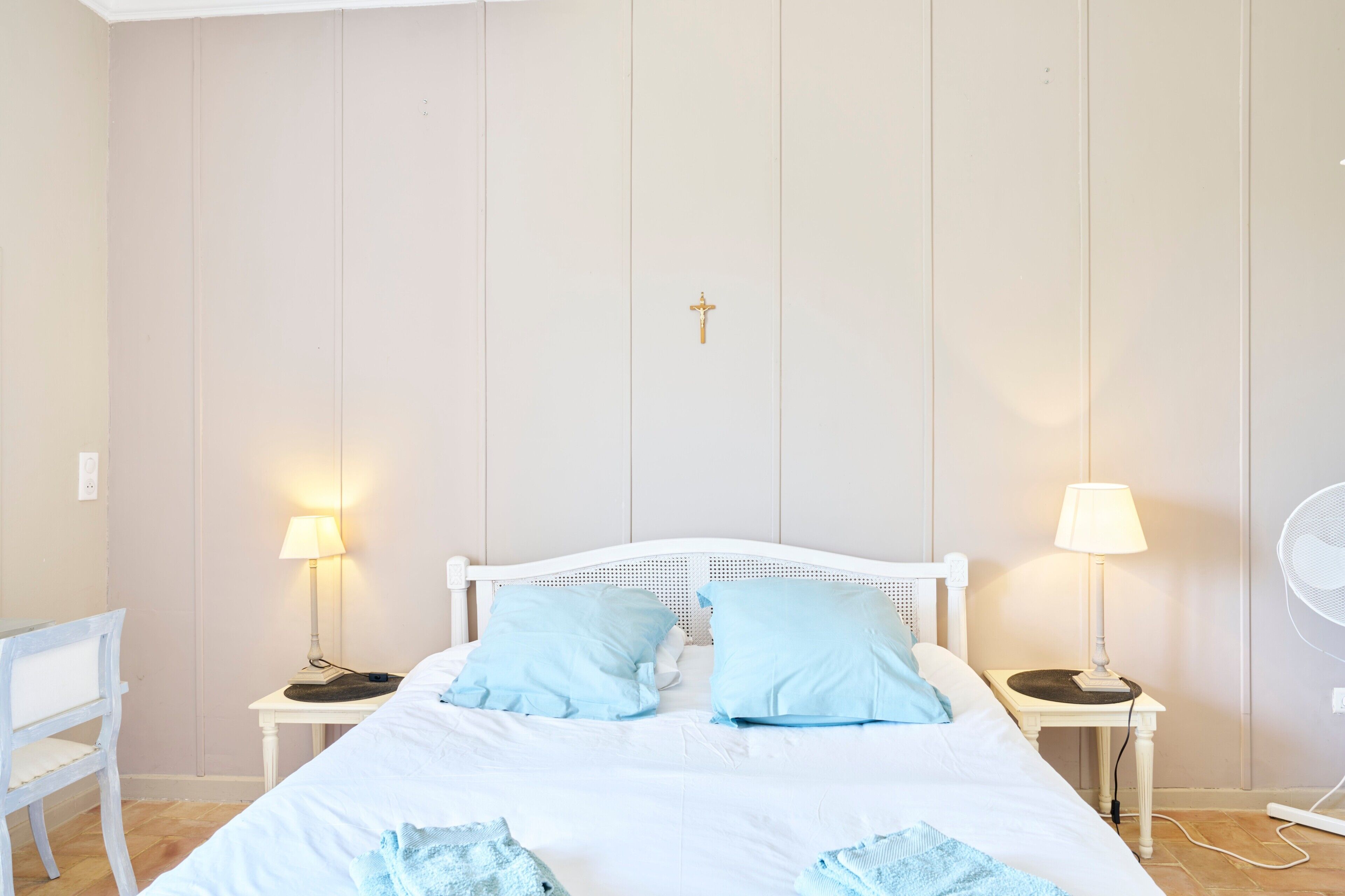 A bedroom with a white bed, two bedside tables with lamps, a white chair, and a wall-mounted fan.