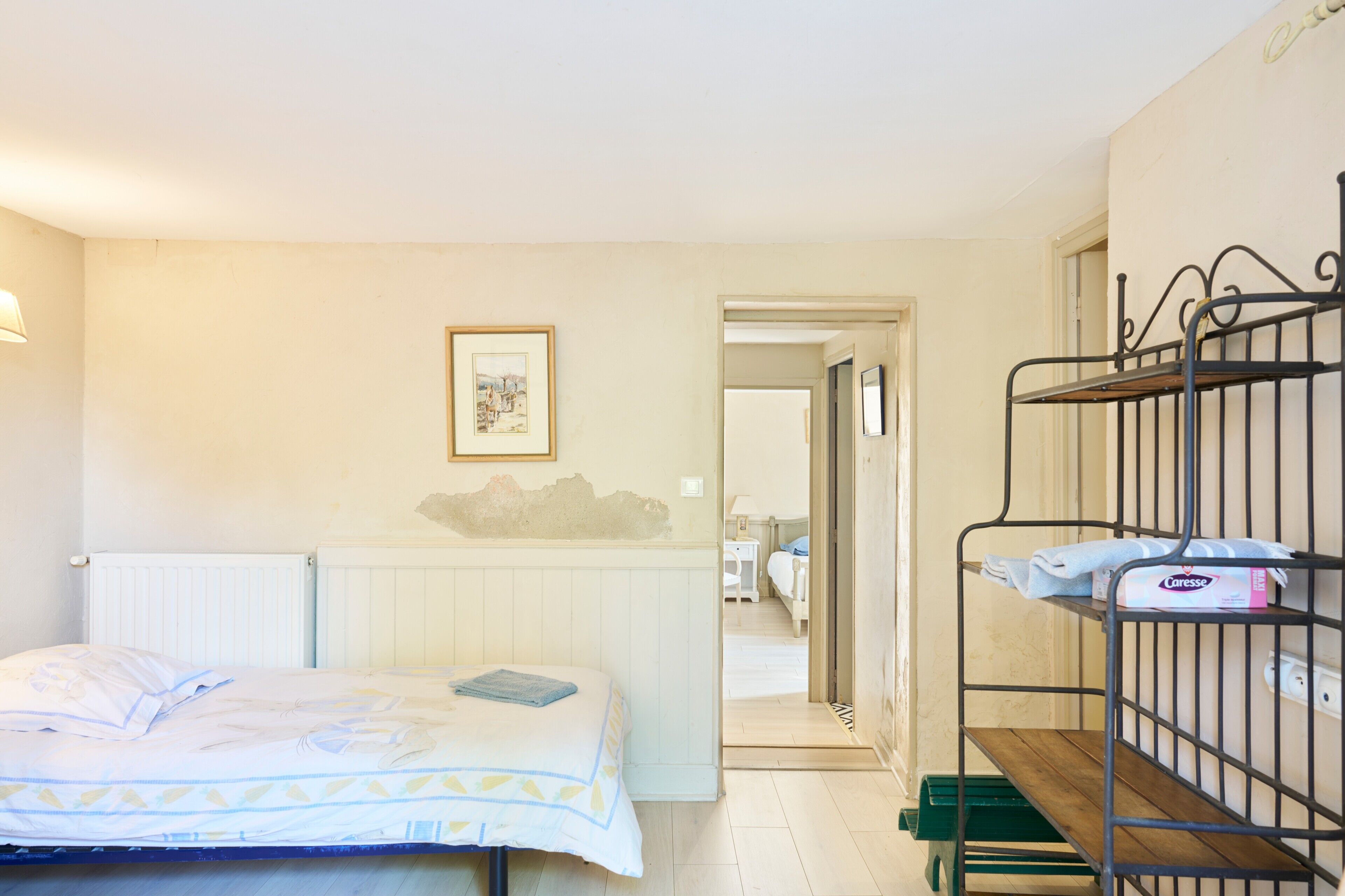 Una habitación con una cama, un radiador, un cuadro en la pared y una litera con una toalla y una caja.