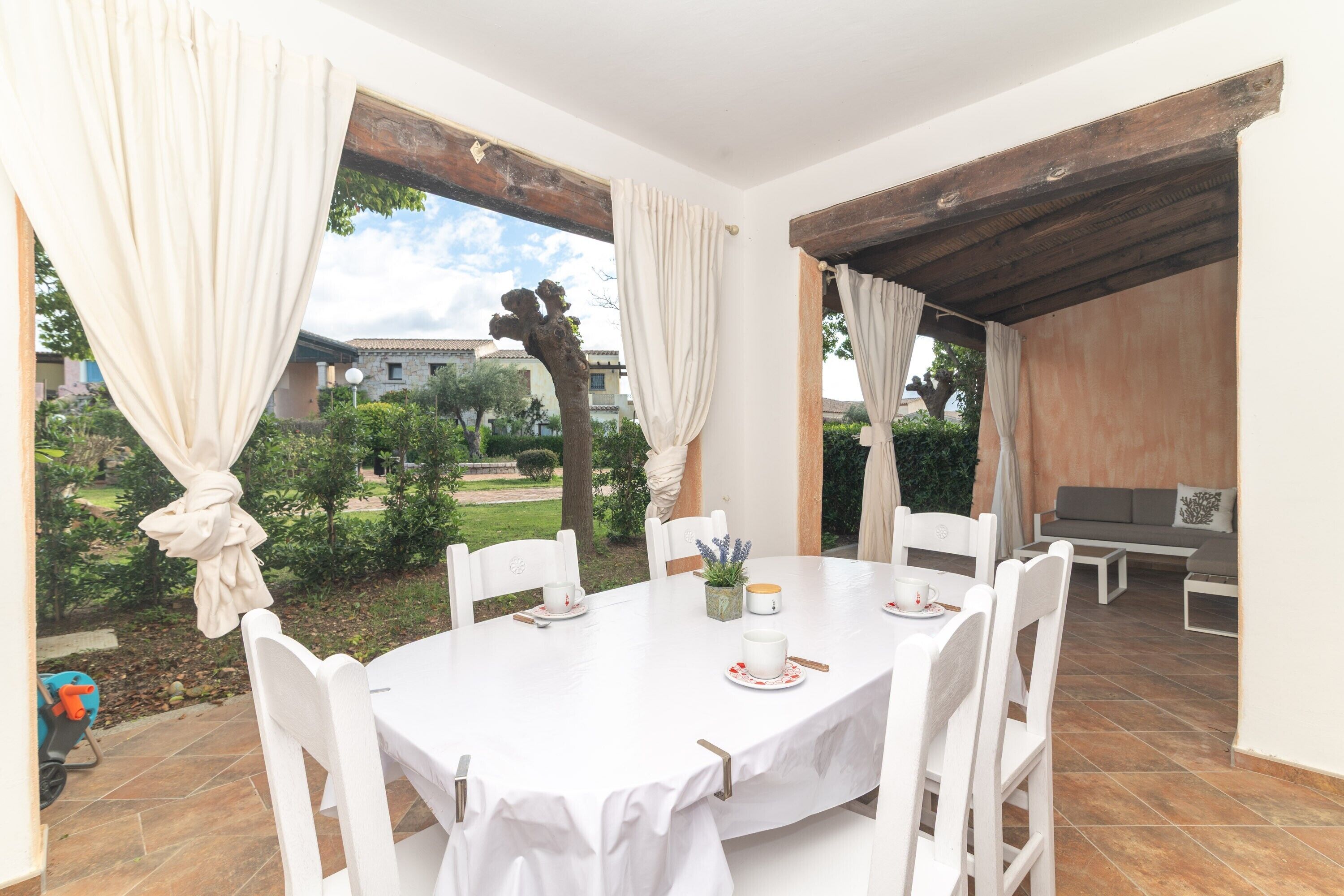 Una mesa redonda con sillas blancas, un mantel blanco y una vista de un jardín y edificios al fondo.