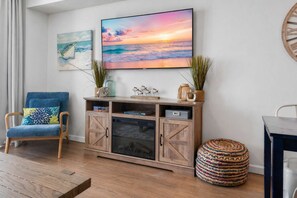 A living room with a TV, wooden cabinet, blue armchair, and a round woven ottoman.