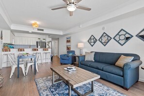 A modern living room with a blue sofa, a wooden coffee table, and a dining area with white chairs.