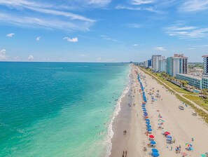 A beach with a long stretch of sand, a pier in the distance, and high-rise buildings along the shore.