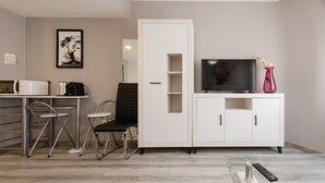 Ein modernes Wohnzimmer mit Flachbildfernseher, weißem Schrank, Esstisch mit Stühlen und einem gerahmten Kunstwerk an der Wand.