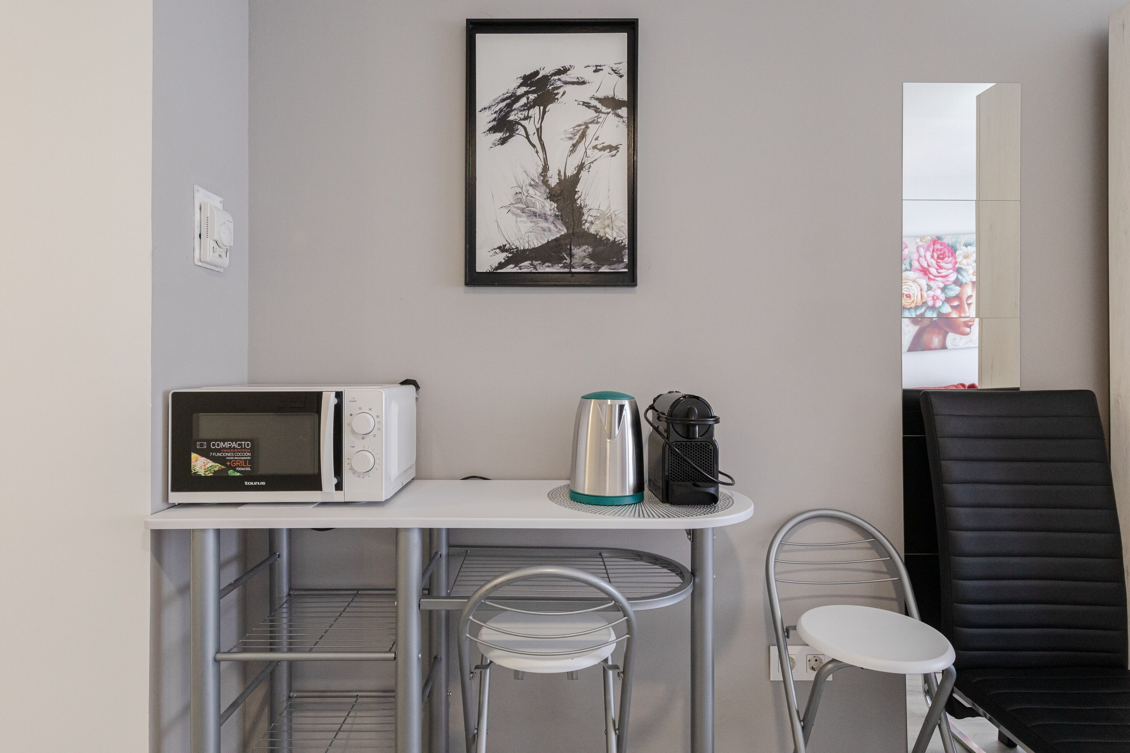 Eine kompakte Küchenzeile mit Mikrowelle, Wasserkocher und Kaffeemaschine, ein kleiner Tisch mit Stühlen und ein gerahmtes Kunstwerk an der Wand.