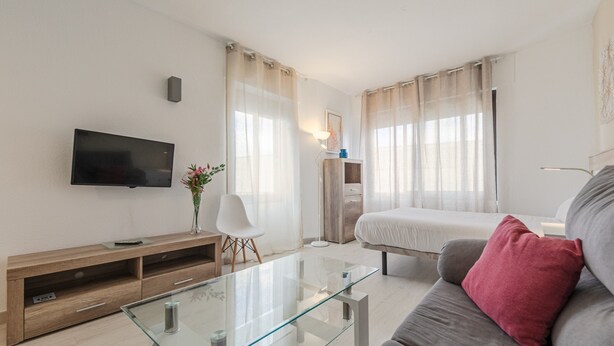 Una sala de estar moderna con una mesa de centro de vidrio, un sofá, un televisor montado en la pared, un mueble de entretenimiento de madera y una cama con cortinas.