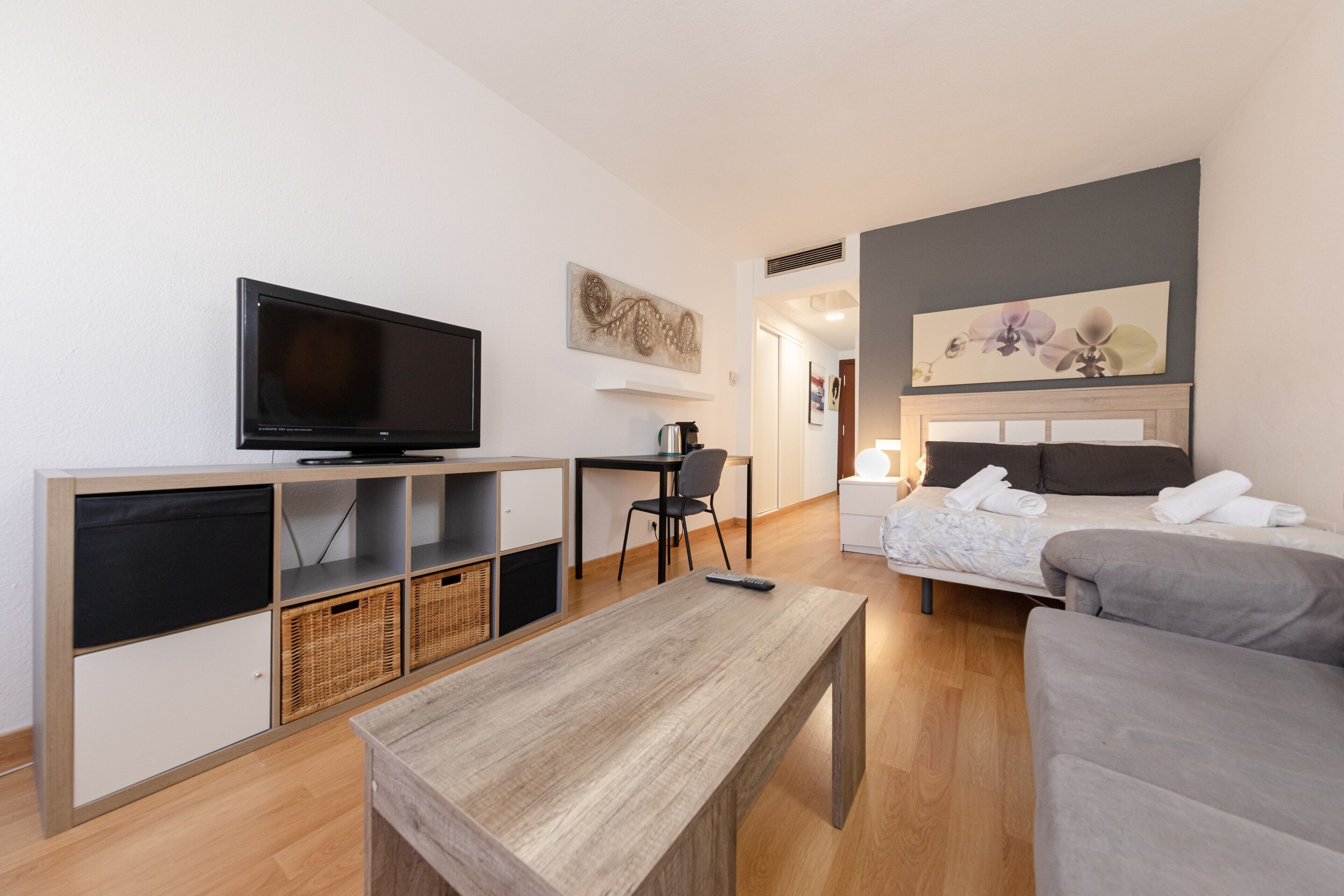 Une chambre d’hôtel moderne équipée d’une télévision à écran plat, d’un bureau en bois, d’un canapé gris, d’un lit avec une tête de lit et d’une œuvre d’art accrochée au mur.