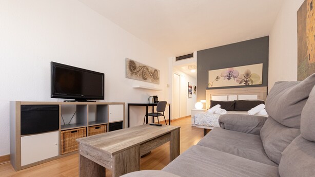 A modern living room with a flat-screen TV, a wooden coffee table, a grey sofa, a dining area with a chair, and a kitchen area with a kettle. - Apartment 'Chick' with Wi-Fi and Air Conditioning (Sevilla)