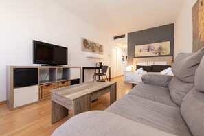 A modern living room with a flat-screen TV, a wooden coffee table, a grey sofa, a dining area with a chair, and a kitchen area with a kettle.