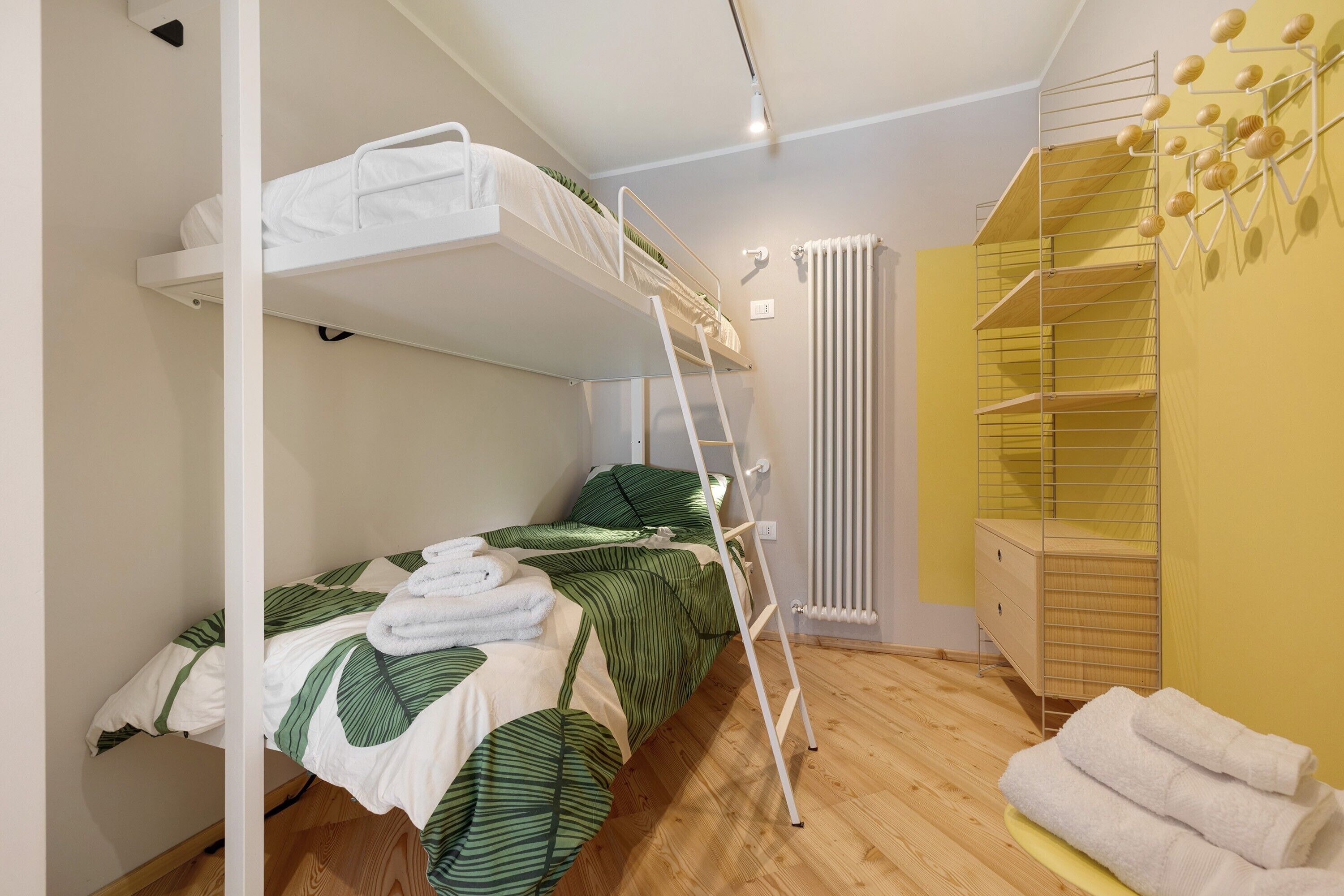 Un lit superposé avec une échelle, une table de chevet, un radiateur et un mur jaune avec une étagère.