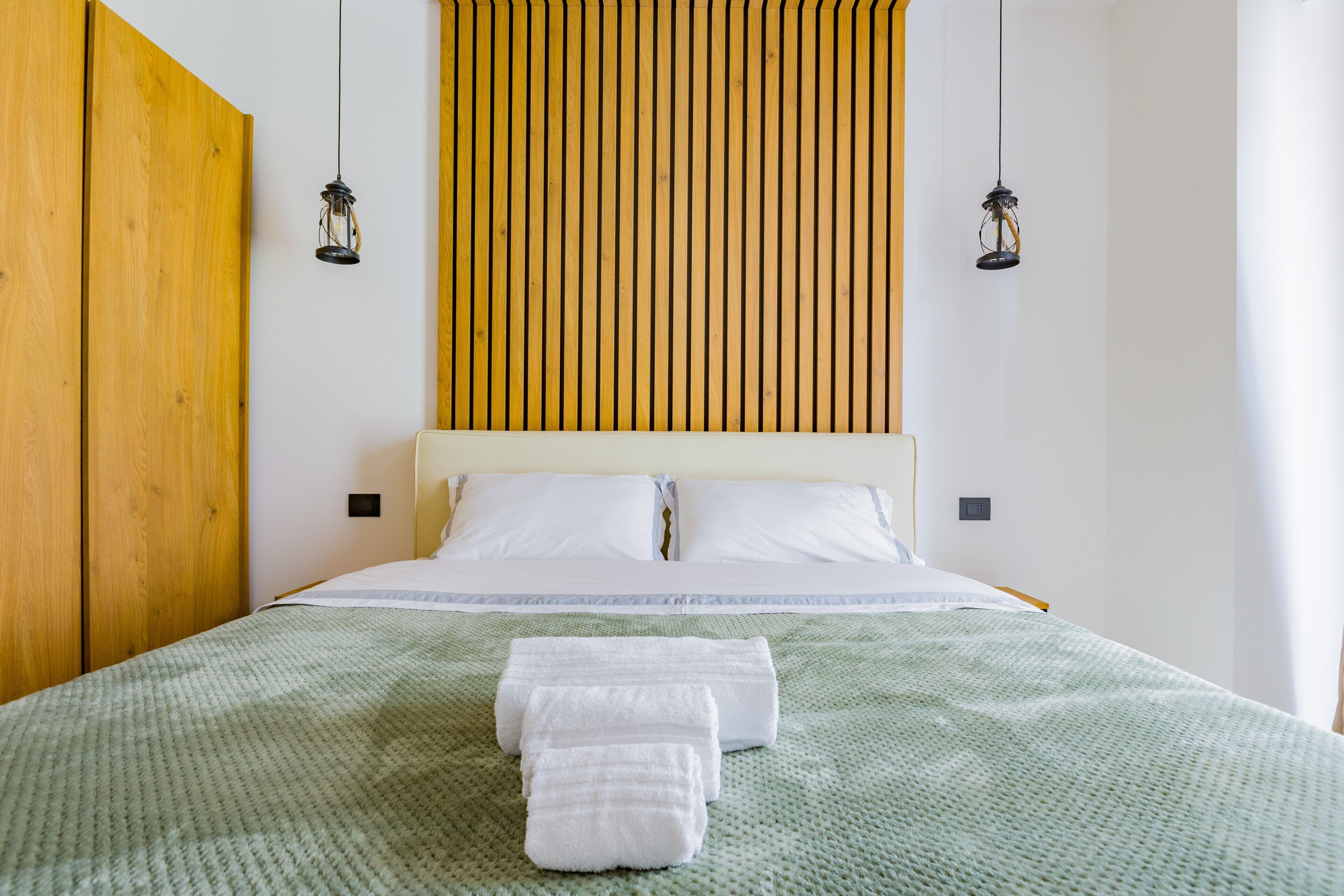 Ein ordentlich bezogenes Bett mit weißen Leinen und einer grünen Decke, Holzvertäfelung an der Wand und zwei hängende Pendelleuchten.