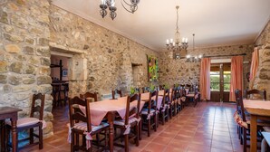 Comedor con paredes de piedra, mesas y sillas de madera, lámpara de araña y vistas a través de una puerta.