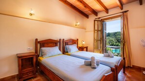 Habitación doble con vigas de madera, dos camas, mesitas de noche, un ventanal con cortinas y vistas a la vegetación.
