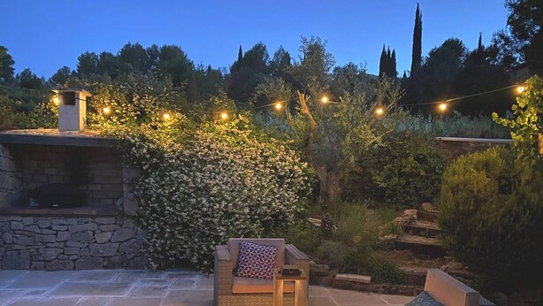 Un área de patio al aire libre con suelo de piedra, una barbacoa integrada y mobiliario exterior con cojines y una mesa.