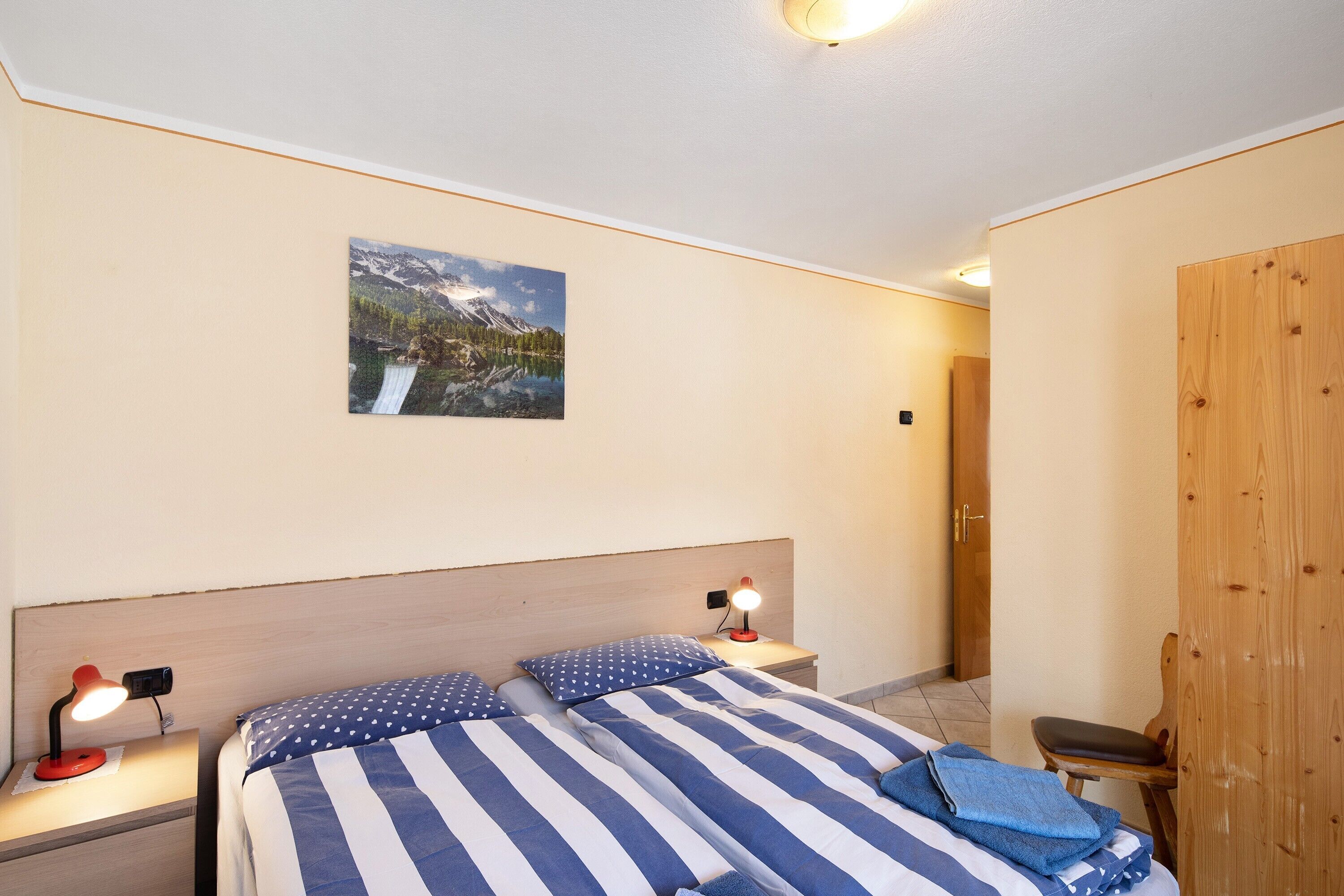A bedroom with a bed, a nightstand, a lamp, a wooden wardrobe, and a painting on the wall.