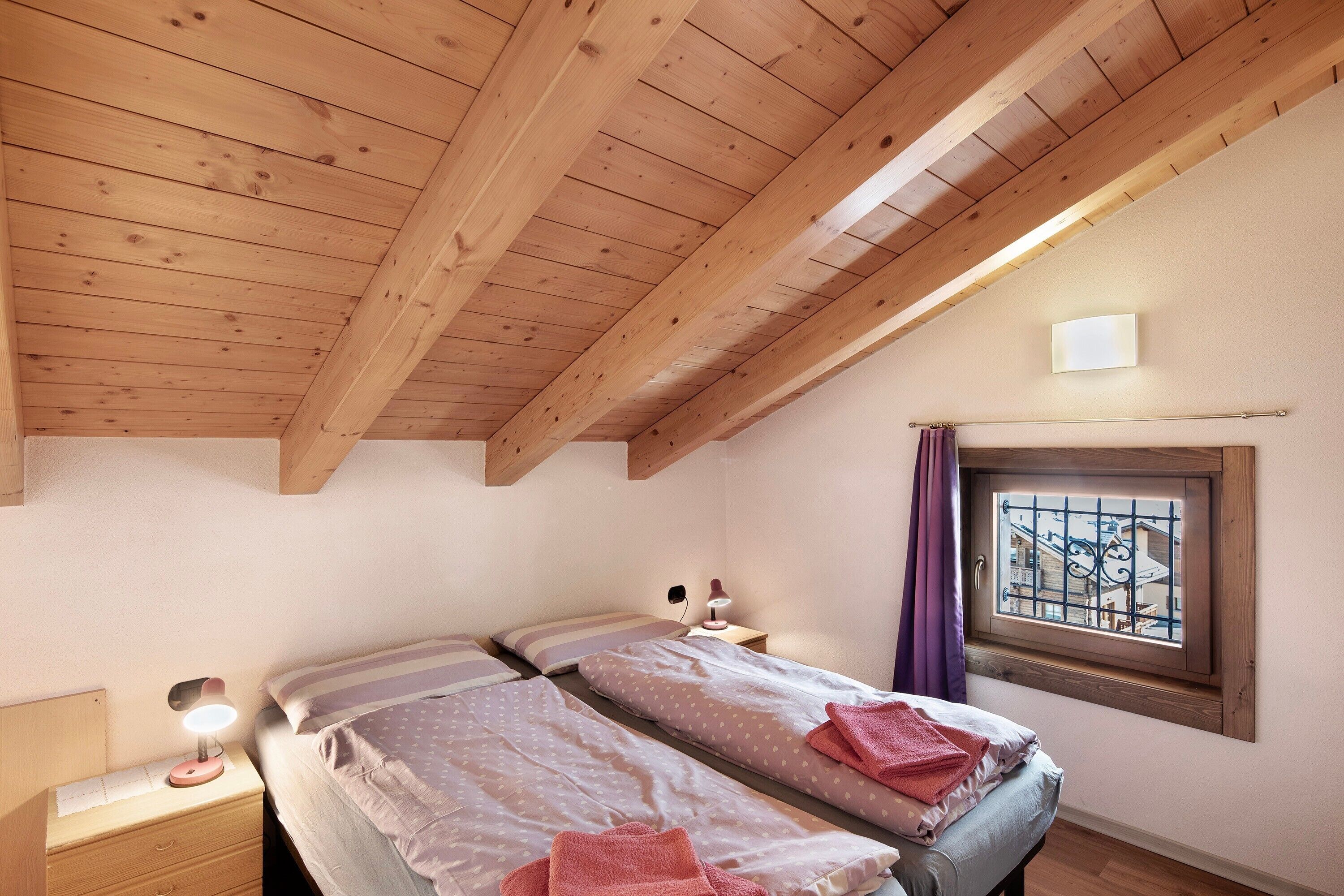 A bedroom with two beds, wooden ceiling beams, a window with bars, and a bedside lamp.