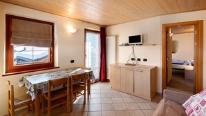 A room with a wooden ceiling, a dining table with chairs, a kitchenette with a TV, and a bed in the adjacent room. - Apartment 'Natale 7' with Mountain View, Balcony and Wi-Fi (Livigno)