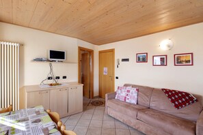 A room with a wooden ceiling, a sofa, a television on a stand, a dining table, and a kitchen area. - Apartment 'Natale 7' with Mountain View, Balcony and Wi-Fi (Livigno)