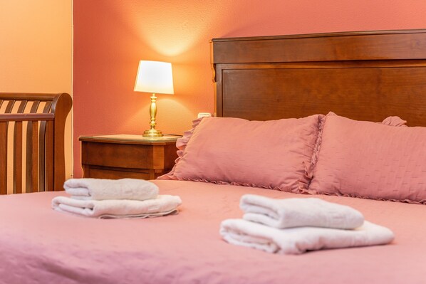Una cama bien hecha con ropa de cama rosa y toallas blancas, una mesita de noche de madera con una lámpara, y una silla de madera.