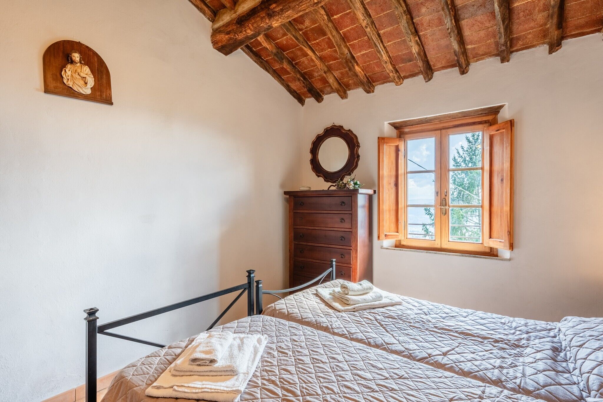 Une chambre avec deux lits, une commode en bois, un miroir et une fenêtre donnant sur des arbres.