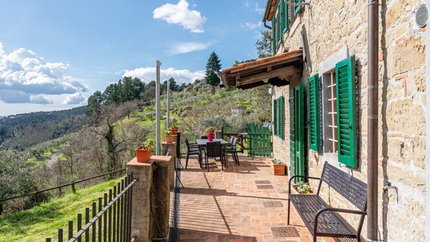 Un edificio de piedra con contraventanas verdes, un patio con mesa y sillas, y vistas a un paisaje montañoso.